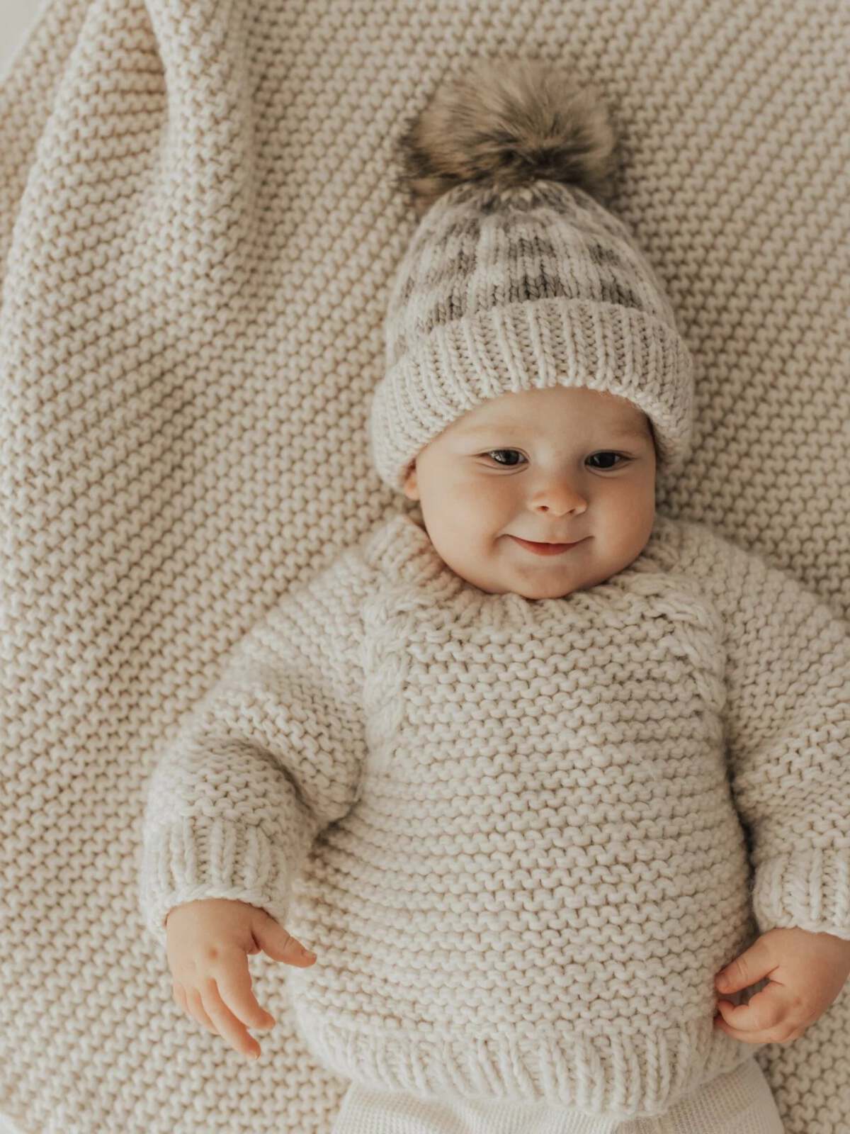 Baby in a cozy knit sweater and hat smiles on a textured blanket. Soft, neutral colors create a warm ambiance.