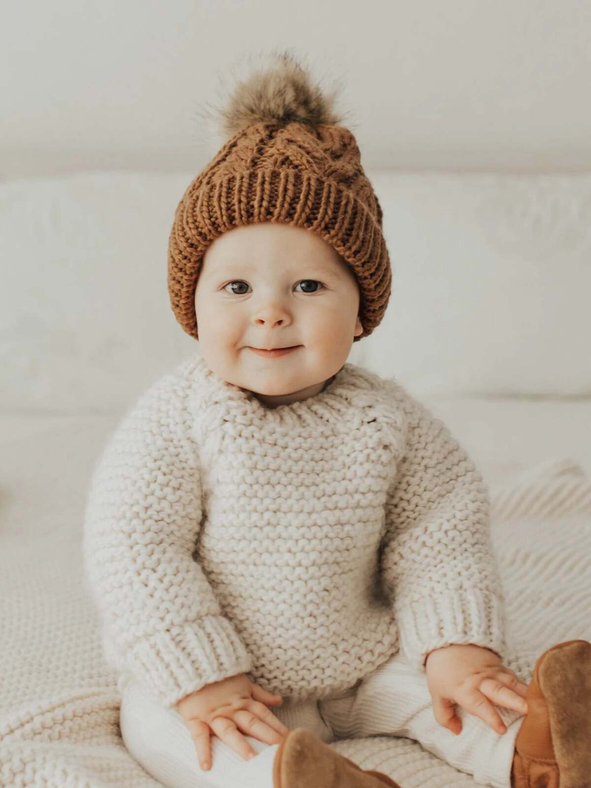 Baby wearing a knitted sweater and brown hat, smiling while sitting on a textured blanket.