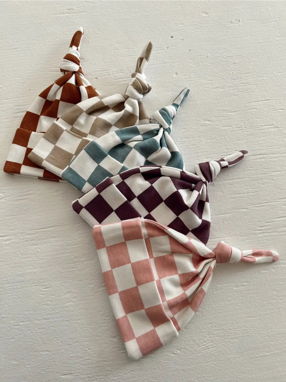 Four checkered baby hats in shades of brown, teal, purple, and pink arranged on a white surface.