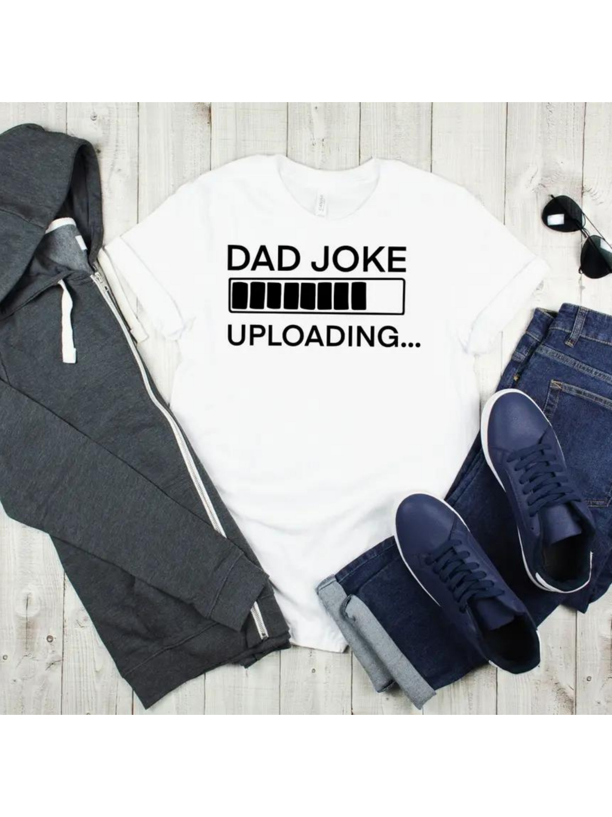 Flat lay of a white t-shirt with "DAD JOKE UPLOADING..." text, gray hoodie, jeans, and sneakers on a wooden surface.