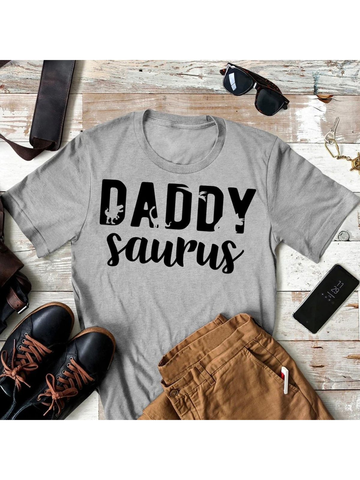 Gray t-shirt with "Daddy Saurus" printed in bold black letters, styled with shoes and accessories.