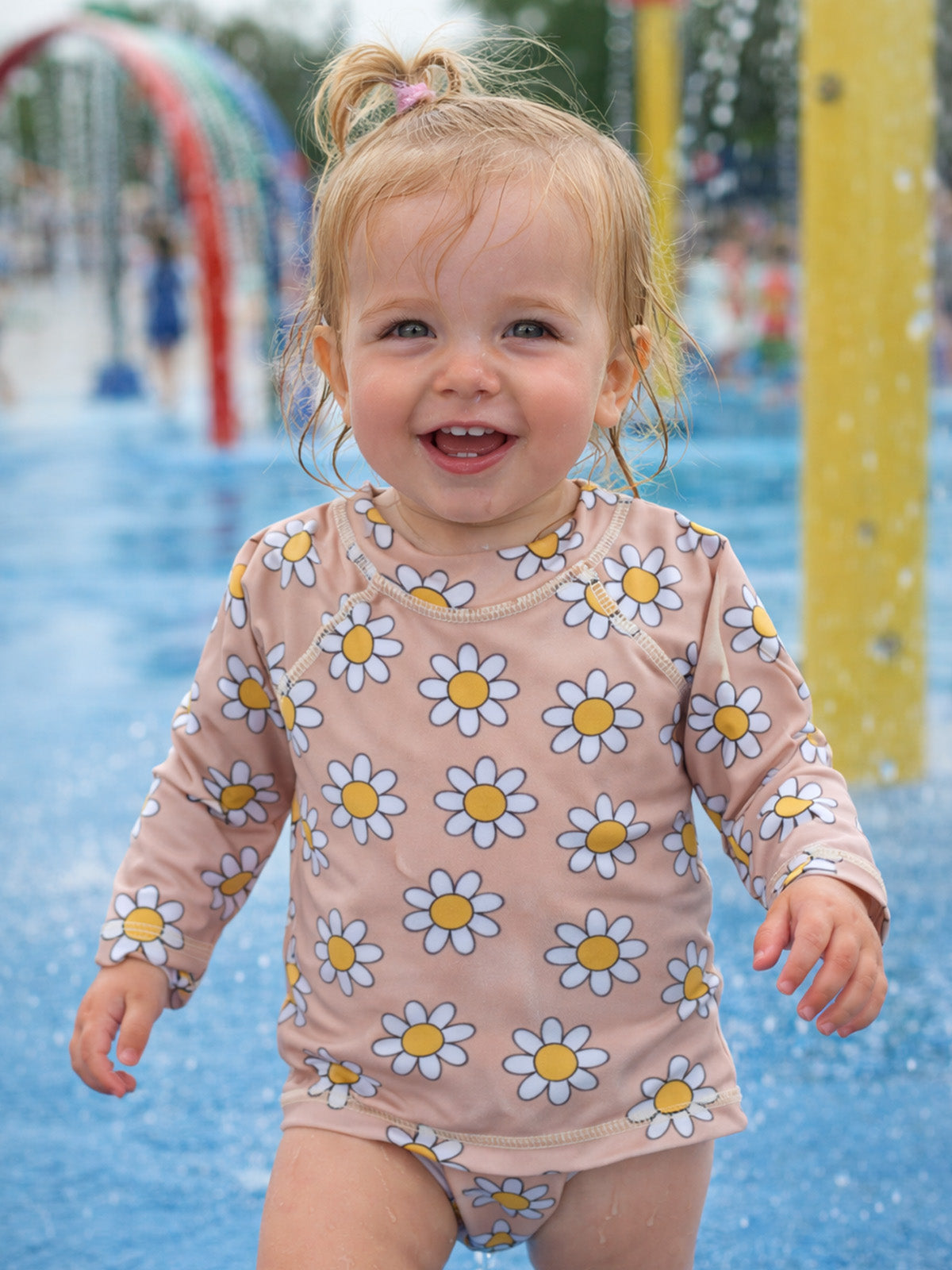 Smiling toddler in a daisy-patterned swimsuit, playfully splashing in water at a colorful splash pad.