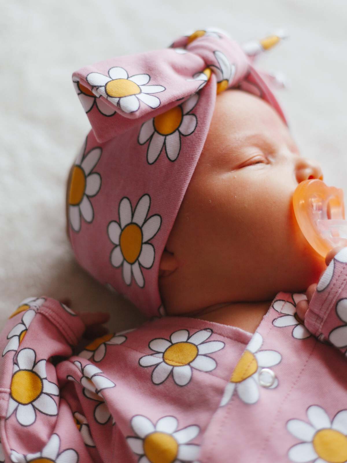 Infant sleeping peacefully, wearing a pink daisy-patterned outfit and a matching headband, with a pacifier.