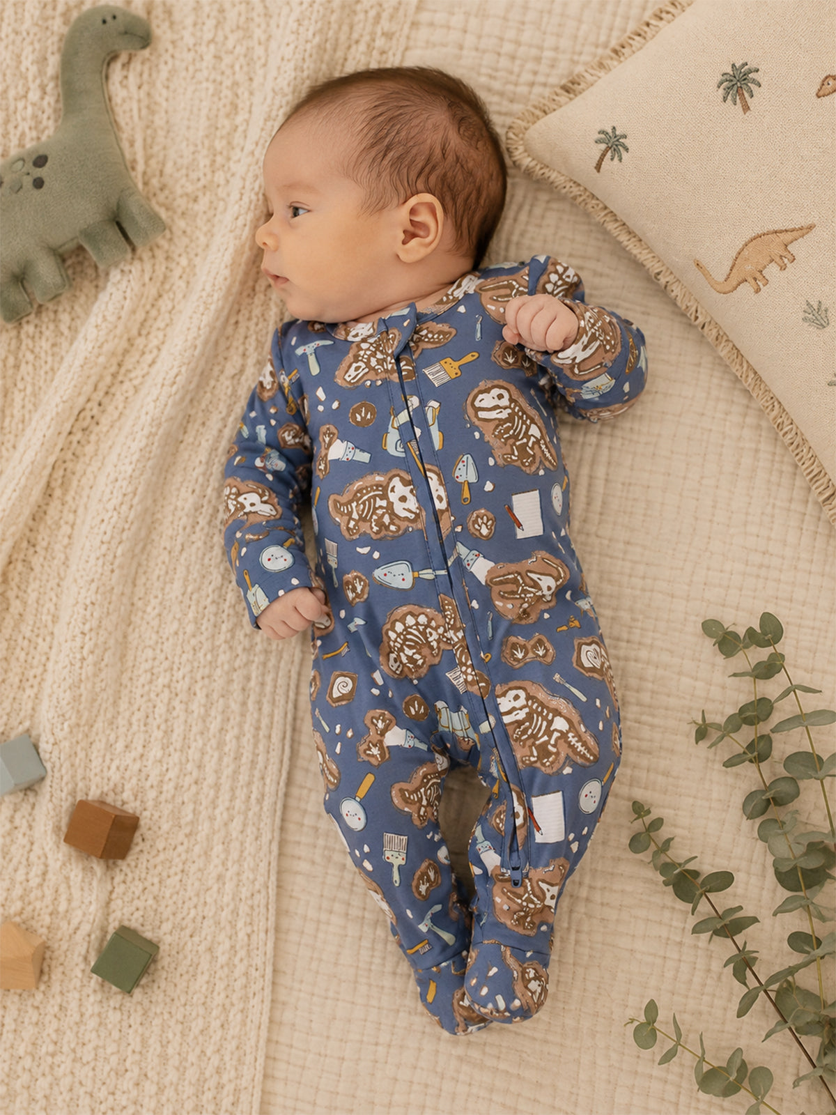 Baby in a blue onesie with animal prints, lying on a textured blanket with toys and greenery nearby.