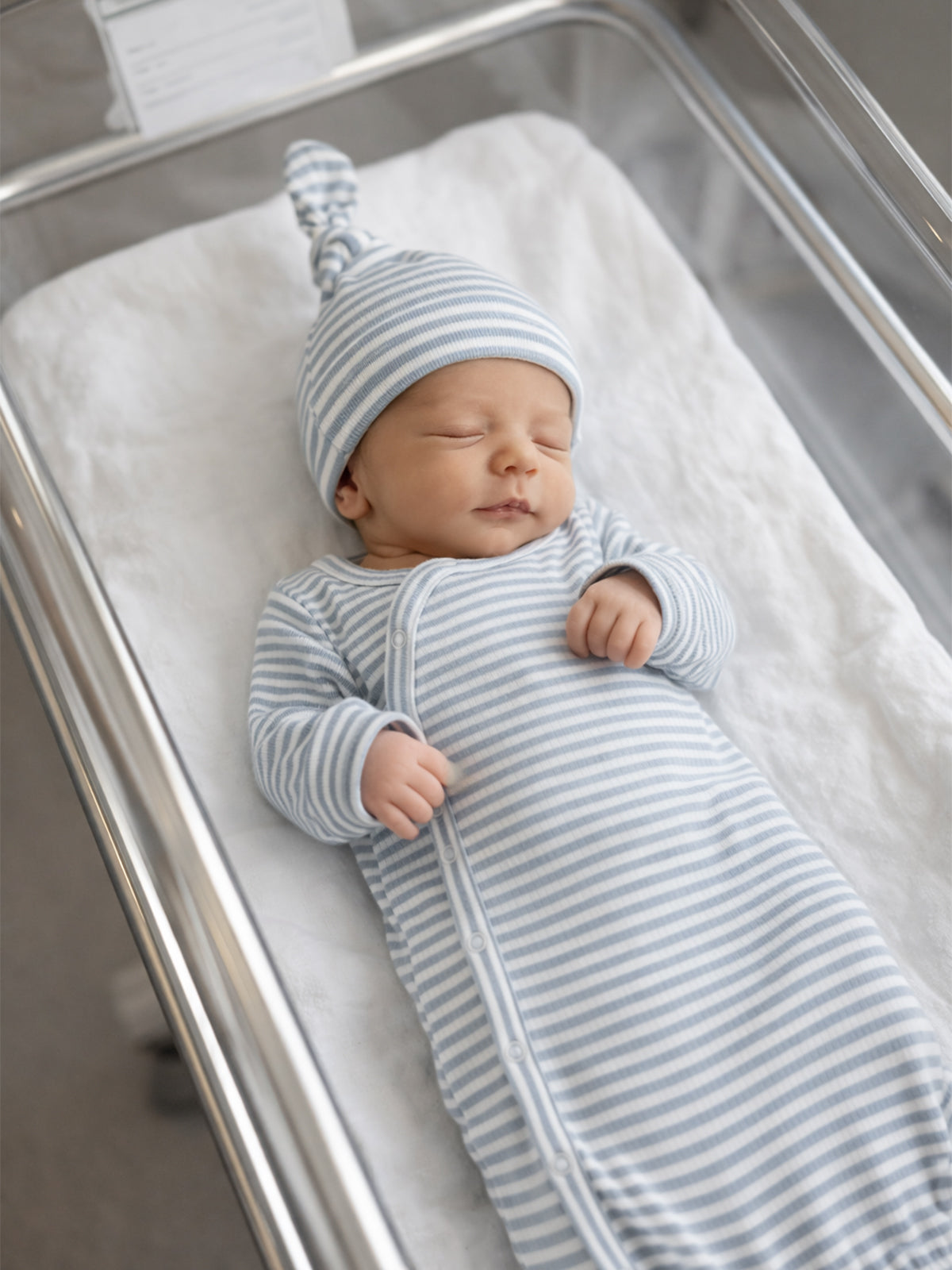 Newborn baby sleeping in a striped blue swaddle and matching hat, in a hospital bassinet.