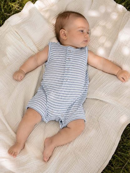 Infant lying on a soft blanket outdoors, wearing a light blue, checkered sleeveless romper.