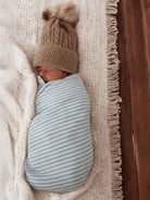 Newborn swaddled in light blue striped blanket, wearing a cozy tan knitted hat on a soft white blanket.