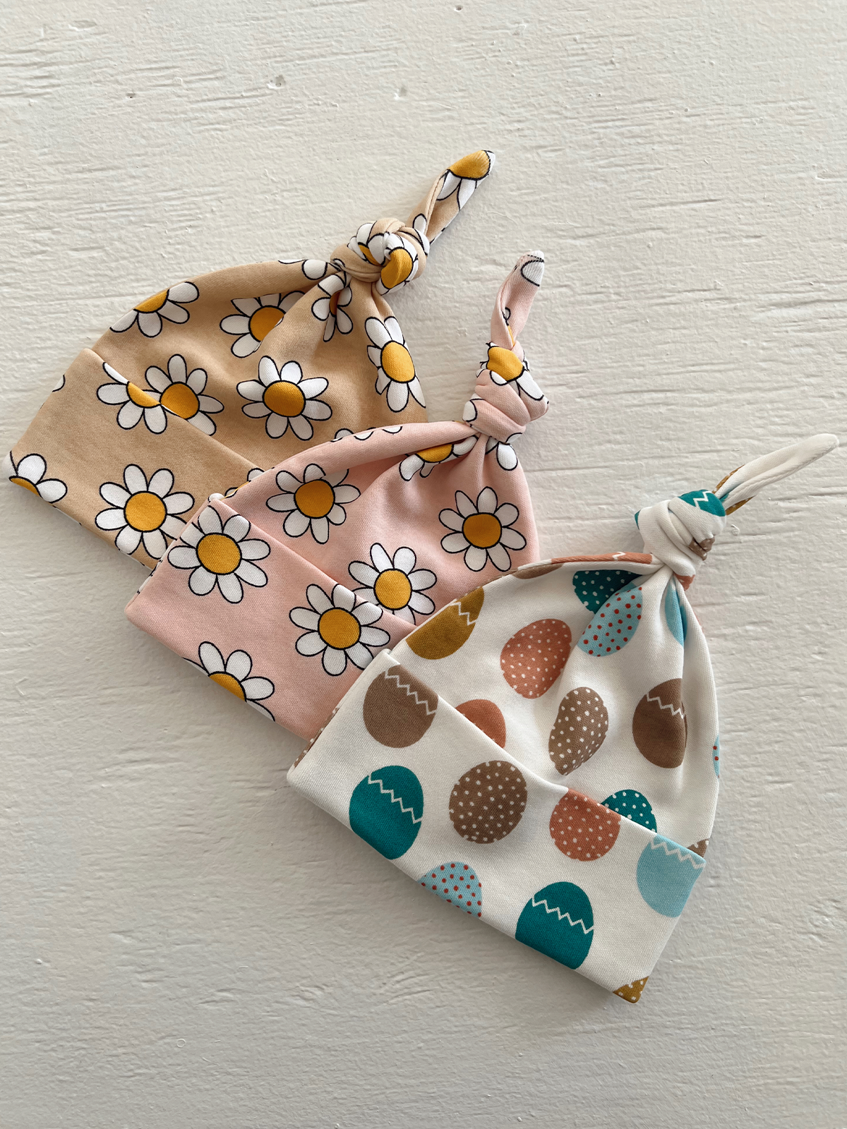 Three baby hats: one with daisies, one pink floral, and one with colorful egg patterns, displayed on a white background.