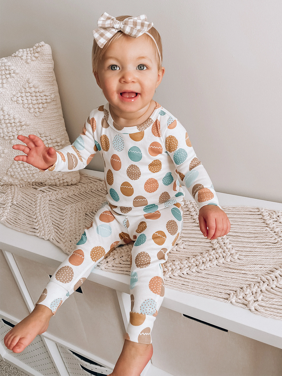 Smiling toddler in patterned pajama set, sitting on a bench with a cozy textured backdrop.