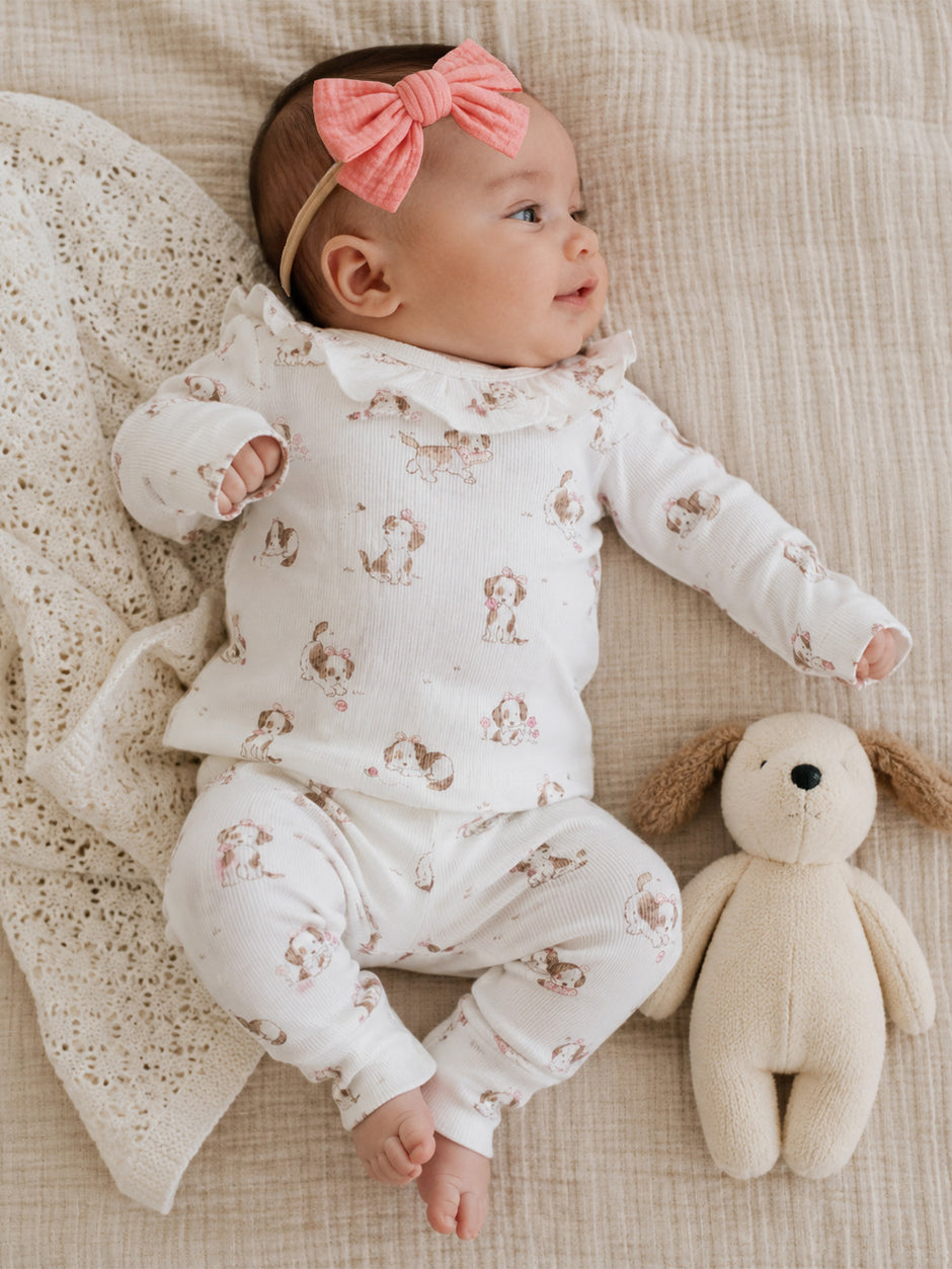 Infant in a patterned outfit with a pink bow, lying next to a soft toy on a textured blanket.
