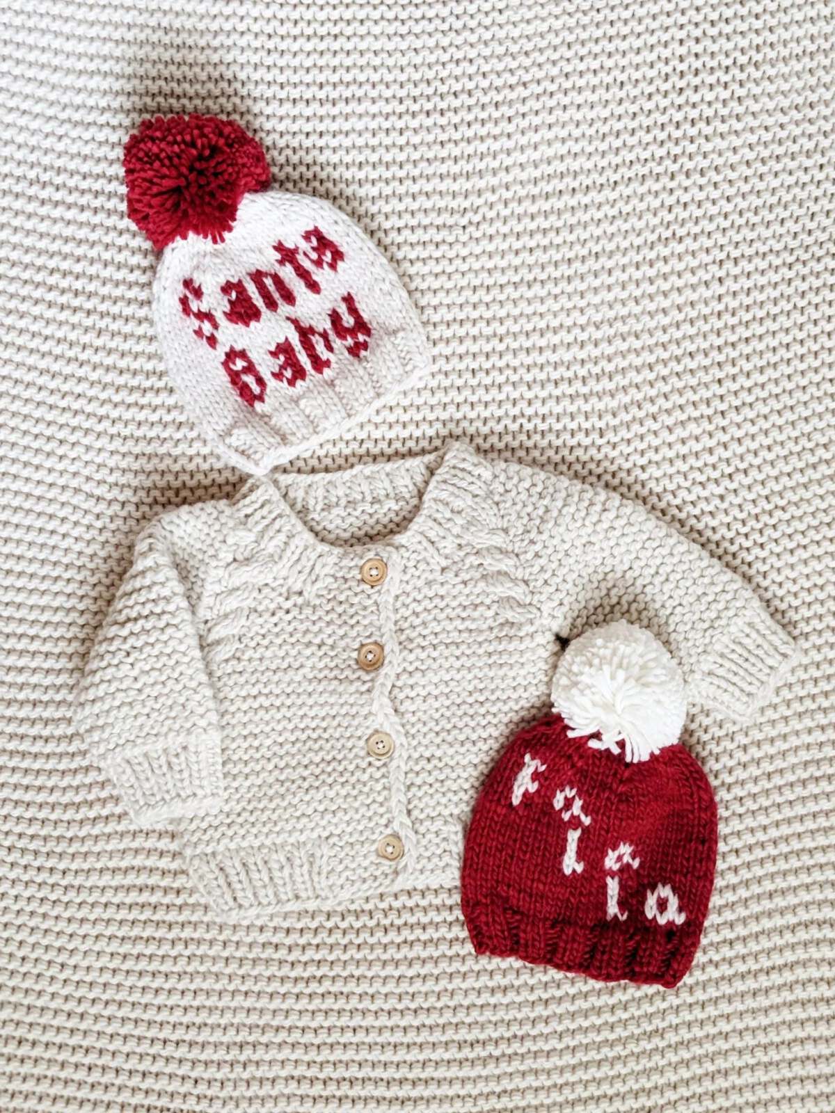 Knit toddler sweater and two beanies on a textured blanket, one saying "Santa Baby" and the other "tia."
