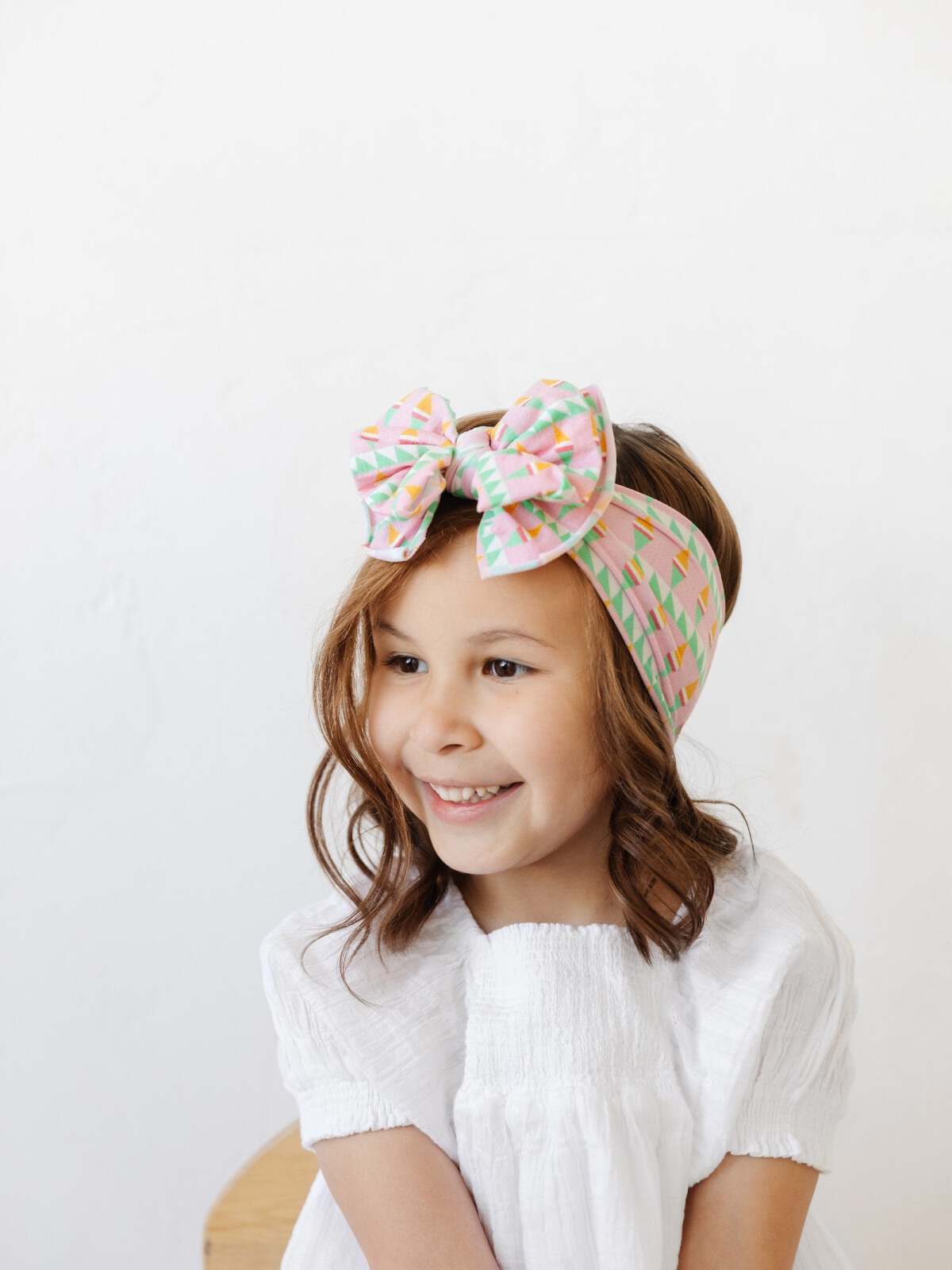 Smiling girl wearing a colorful bow headband and a white shirt, set against a light background.