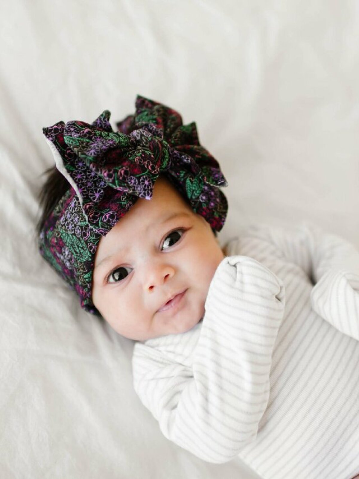 Baby wearing a colorful headband, lying on a light background with a curious expression.