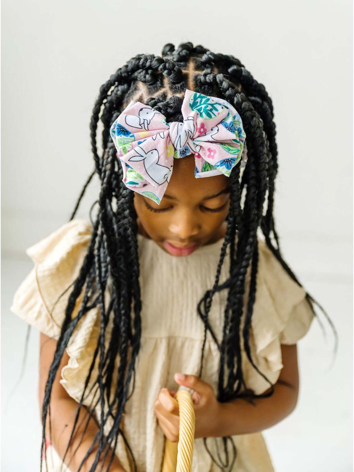 Girl with braids wearing a colorful bow, holding a woven bag, outdoors with a neutral background.