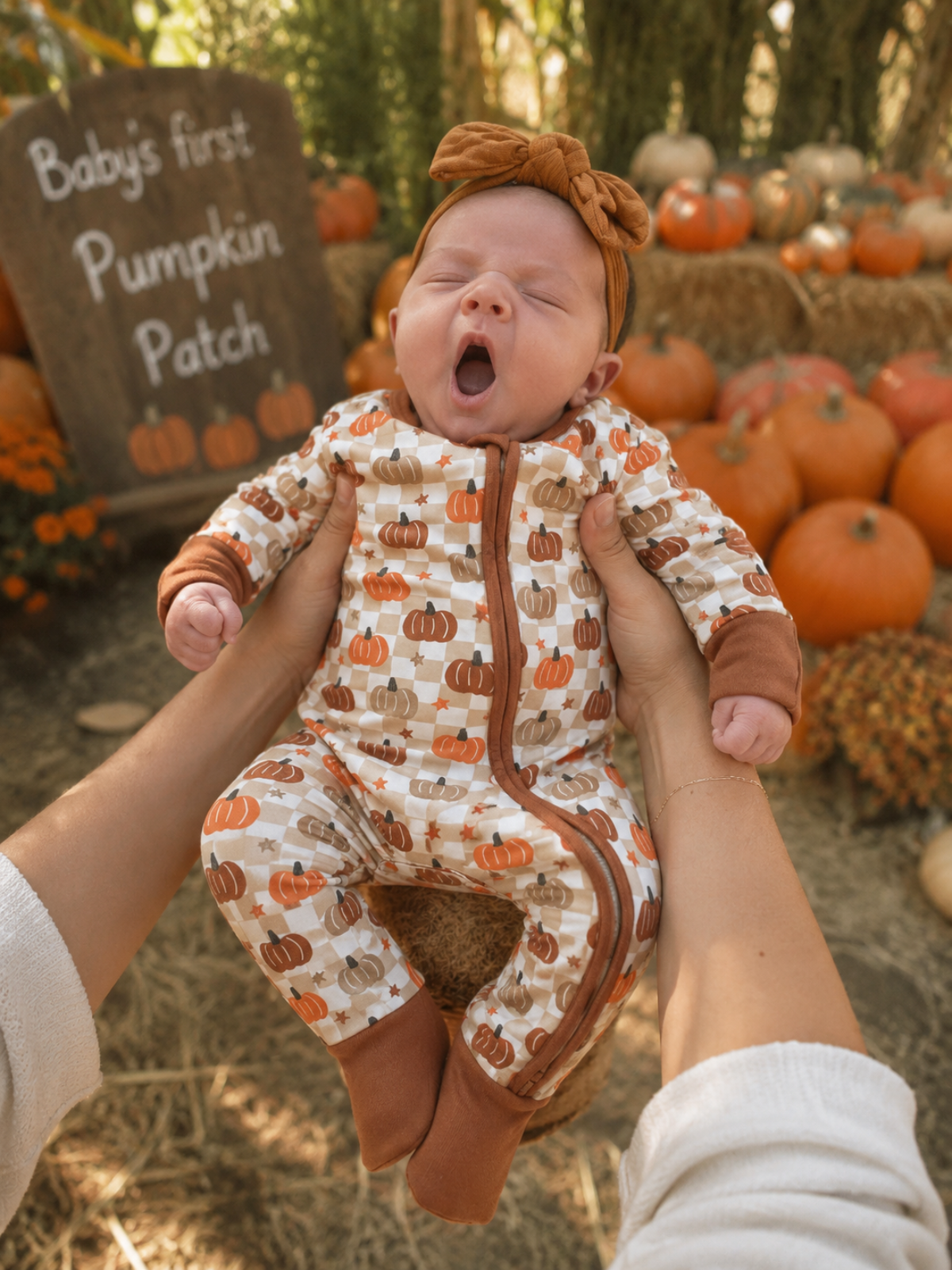 Baby in pumpkin-themed outfit yawning, held up in front of a "Baby's first Pumpkin Patch" sign and pumpkins.