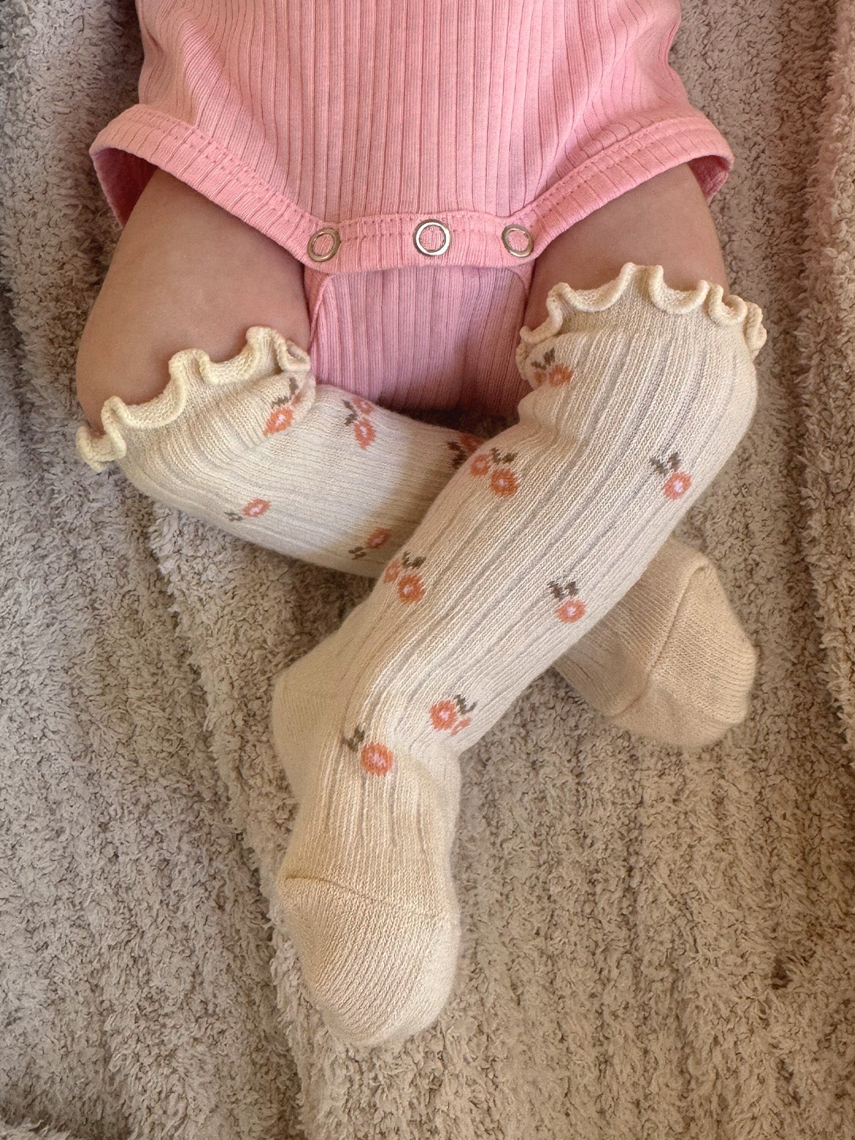 Infant's legs in floral knee-high socks, wearing a pink onesie, resting on a cozy textured blanket.