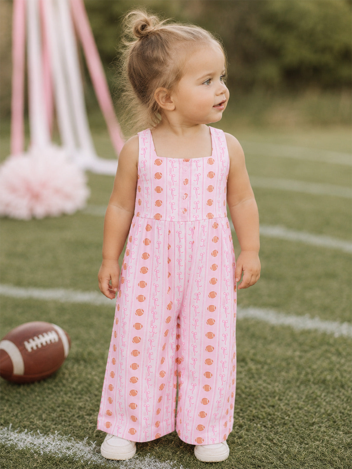 Toddler in a pink patterned jumpsuit stands on grass, smiling with a football nearby.