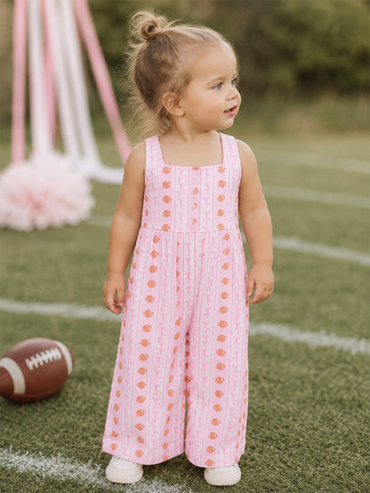 Toddler in a pink patterned jumpsuit stands on grass, smiling with a football nearby.