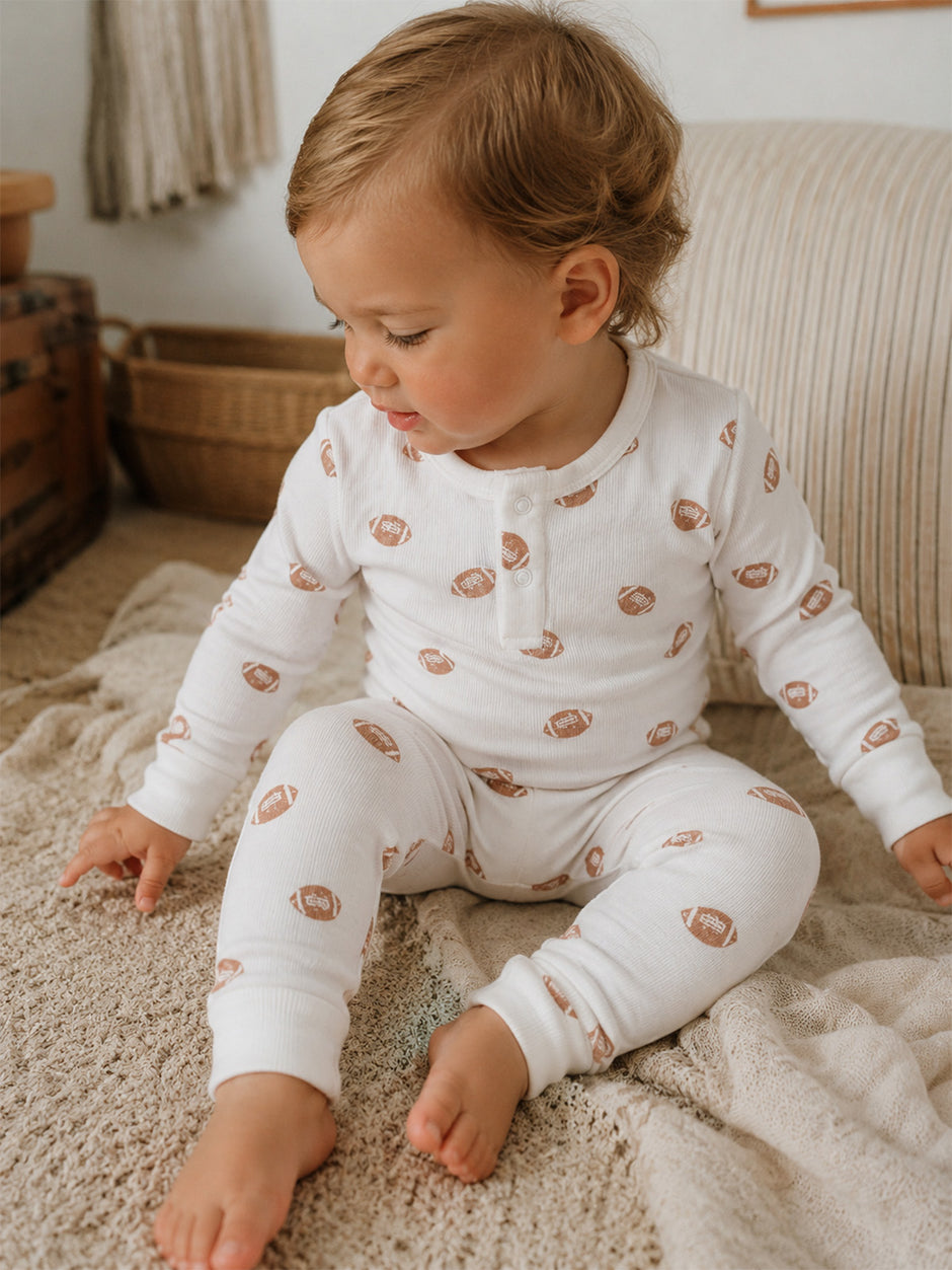 Toddler in white pajamas with football prints, sitting on a cozy blanket, looking thoughtfully to the side.