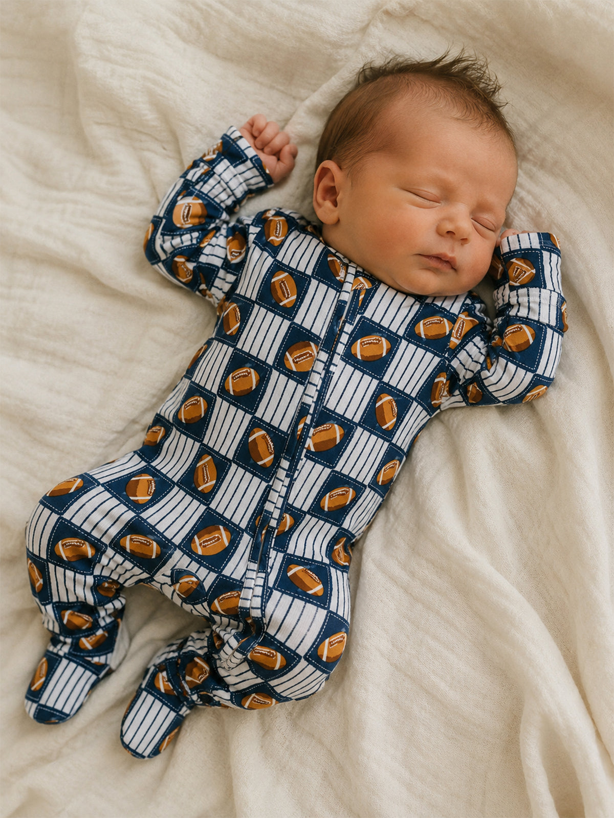 Newborn baby sleeping on a soft blanket, wearing a blue and white football-patterned onesie.