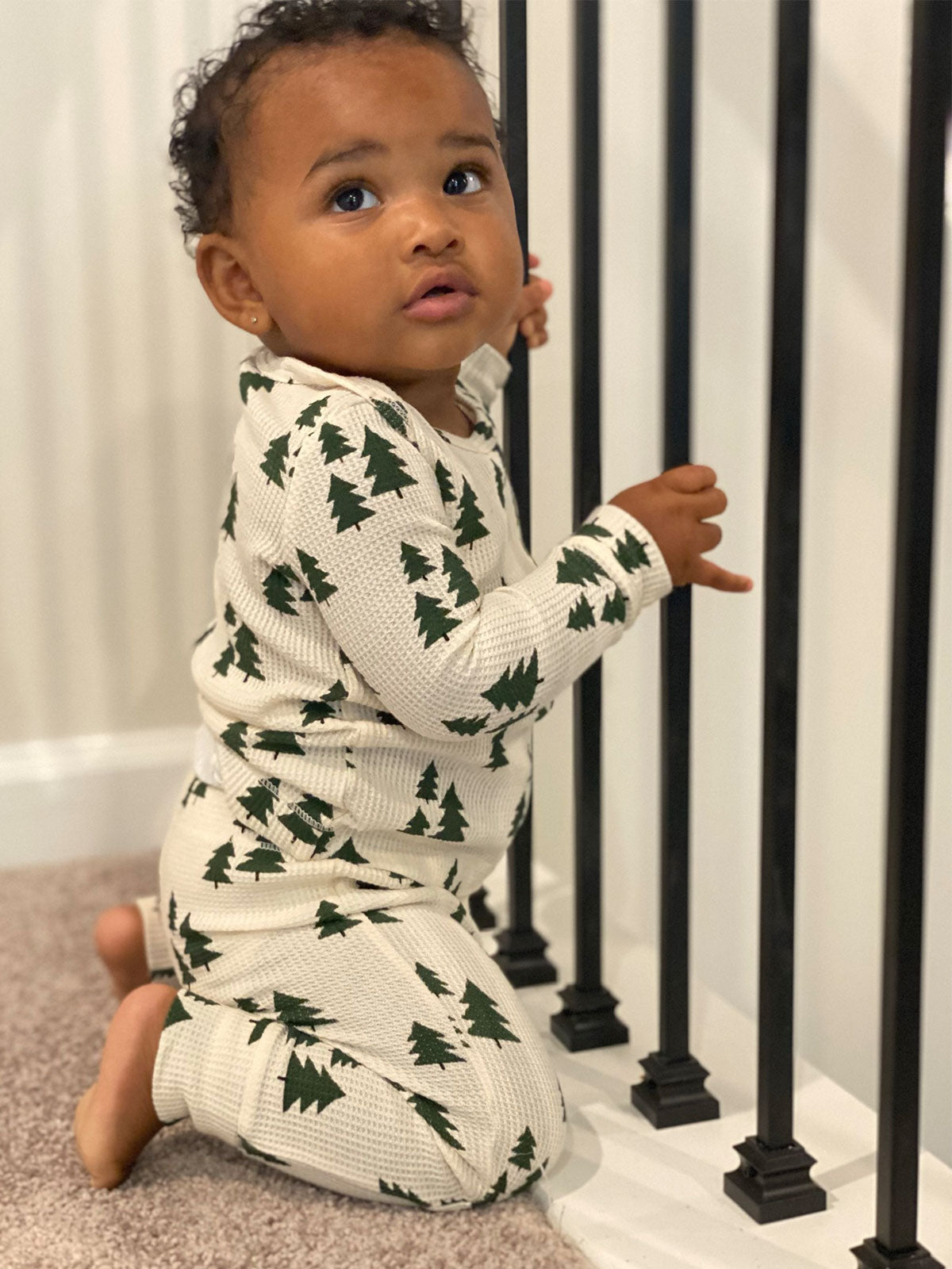Infant in festive pajamas, kneeling by black railing, looking curiously at the camera.