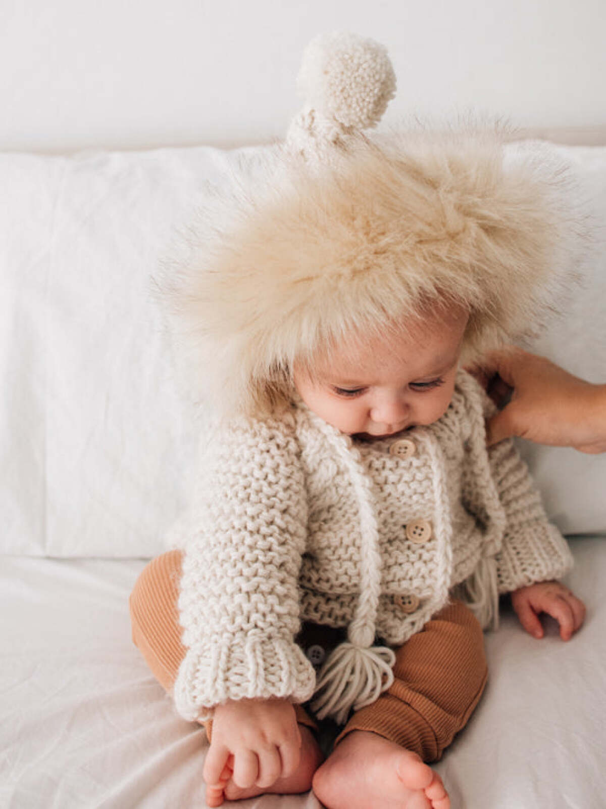 Infant wearing a cozy knit sweater and fluffy hat, sitting on a soft surface, reaching for something.