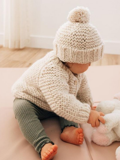Baby in a knitted sweater and hat sits on the floor, playing with a plush toy. Soft, cozy atmosphere.