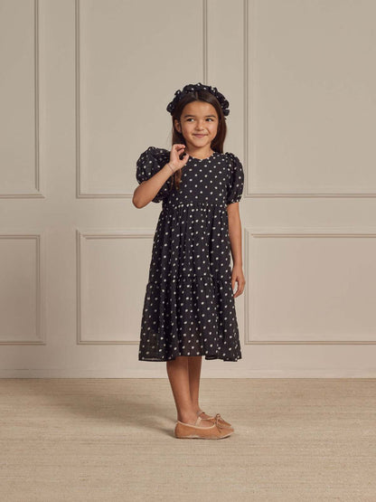 Girl in a polka dot dress and headband smiles while standing against a neutral wall backdrop.