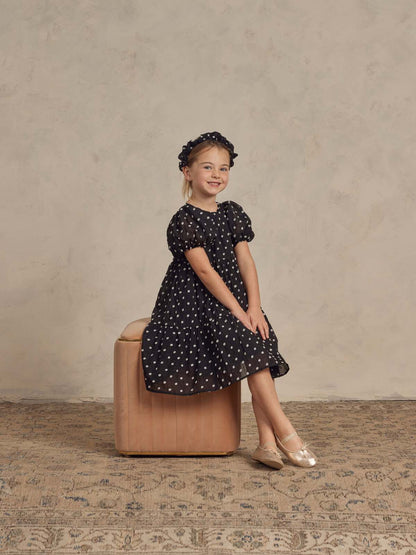 Girl in a polka dot dress and ballet shoes sitting on a beige stool against a textured background.