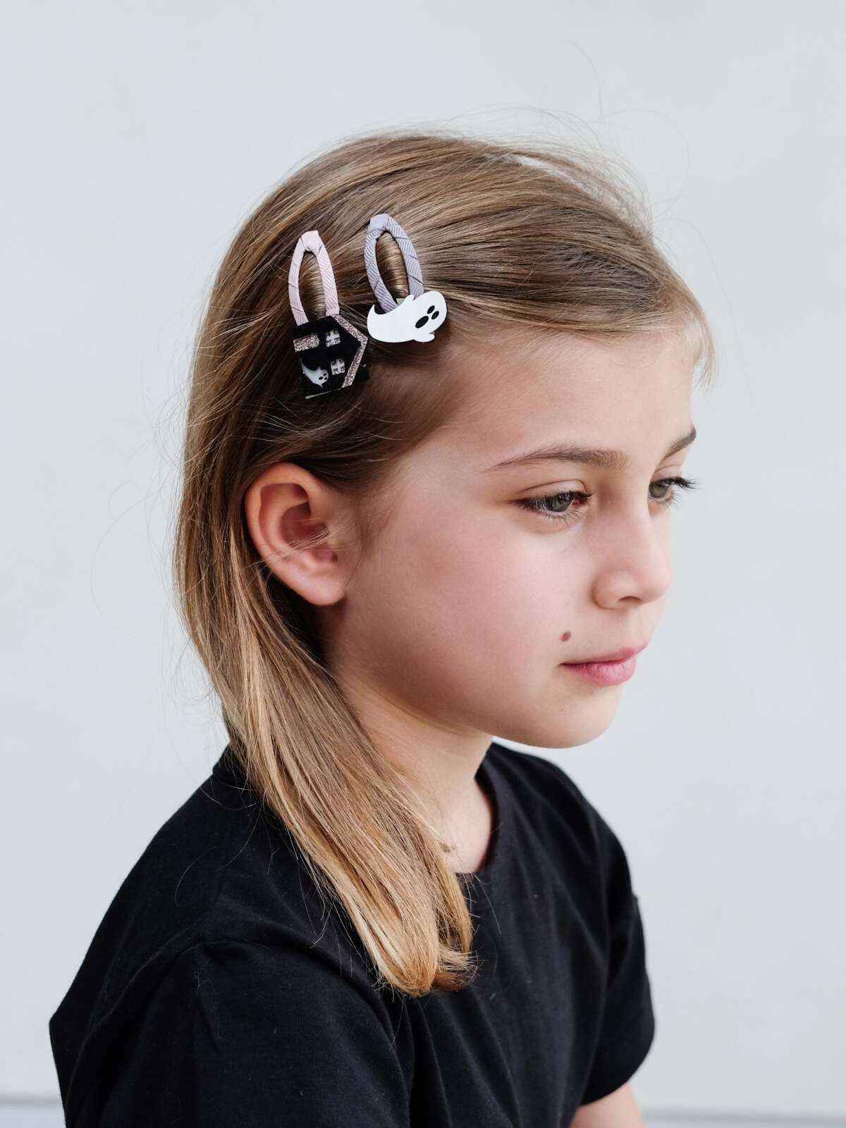 Girl with long hair wearing playful ghost and house-shaped hair clips, looking thoughtfully to the side.