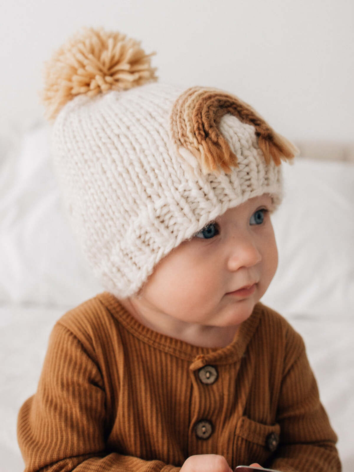 Baby wearing a knotted cream beanie with a pom-pom, sitting on a bed in natural light.