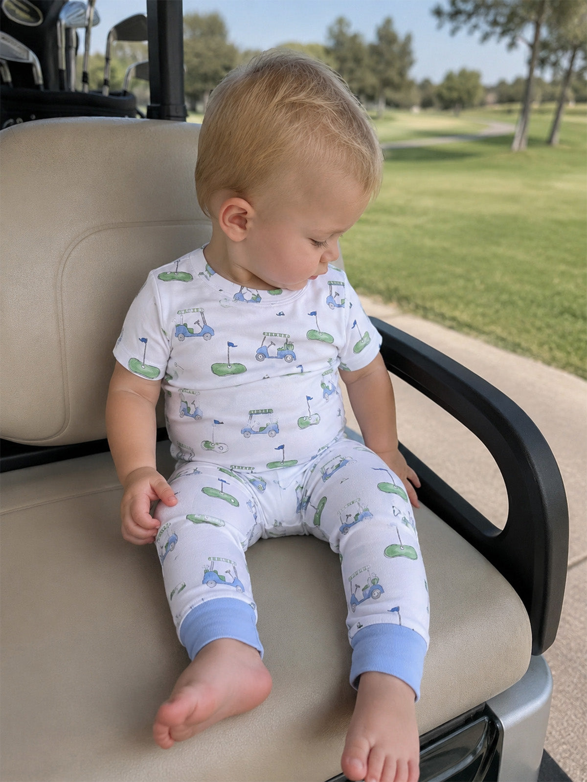 Toddler in golf-themed pajamas sitting on a golf cart seat, with a green landscape in the background.