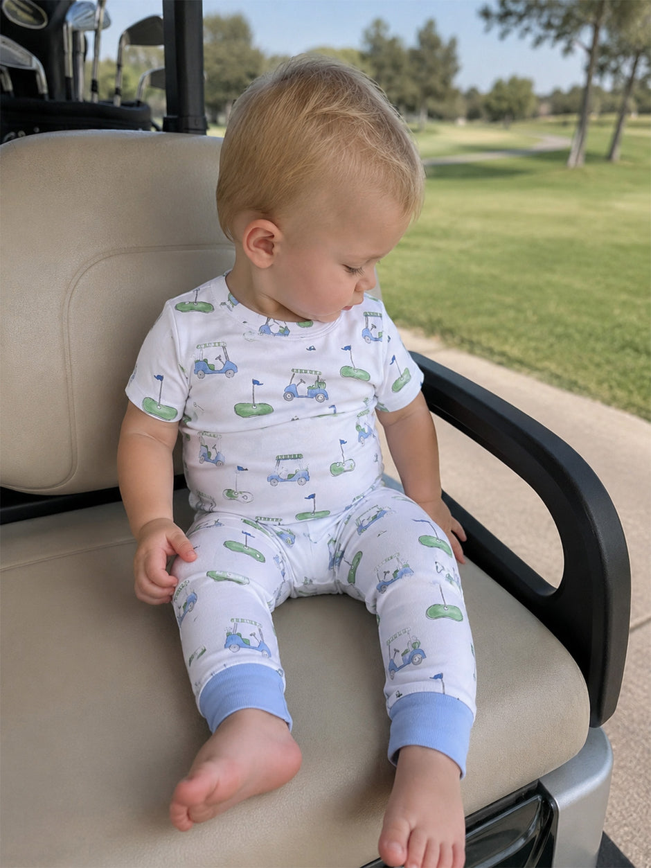 Toddler in golf-themed pajamas sitting on a golf cart seat, with a green landscape in the background.
