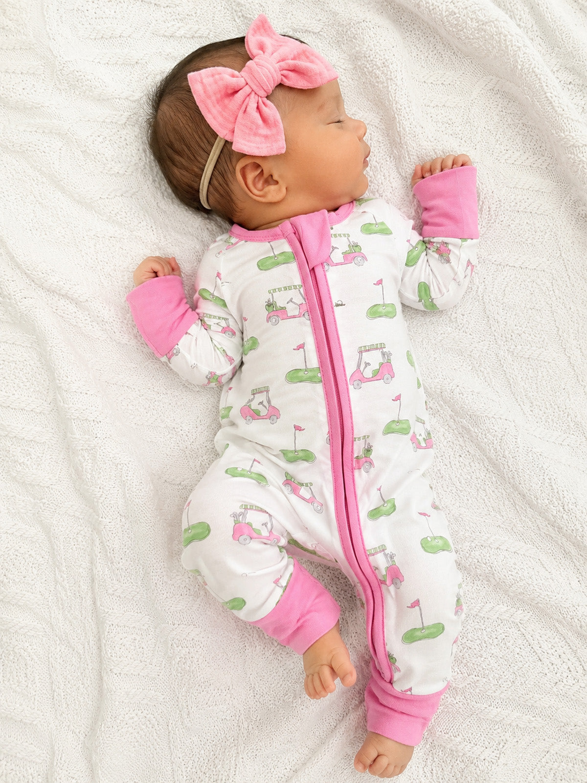 Infant sleeping on a white blanket, wearing a golf-themed onesie and a pink bow headband.
