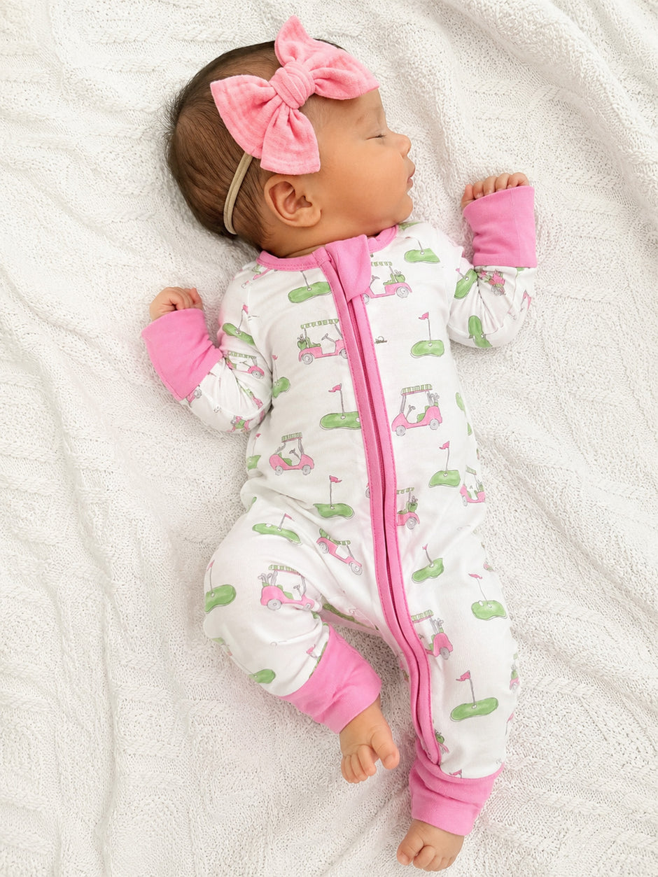 Infant sleeping on a white blanket, wearing a golf-themed onesie and a pink bow headband.
