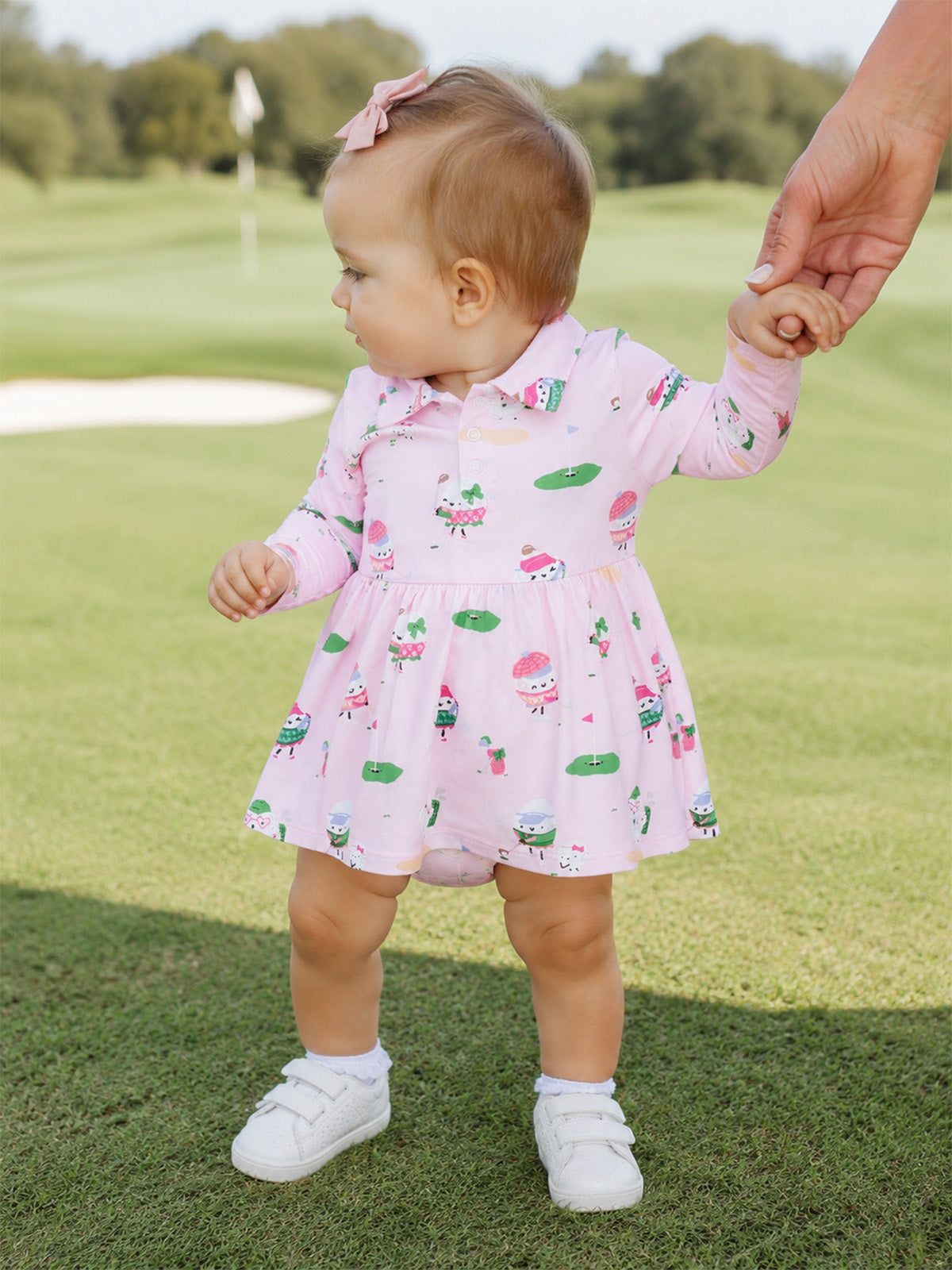 Toddler girl in a pink dress holding an adult’s hand on a golf course, surrounded by greenery.