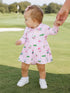 Toddler girl in a pink dress holding an adult’s hand on a golf course, surrounded by greenery.