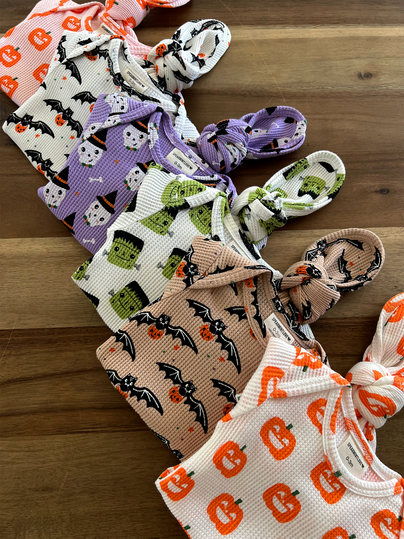 Colorful Halloween-themed baby outfits arranged in a row, featuring bats, pumpkins, and frankenstein designs.