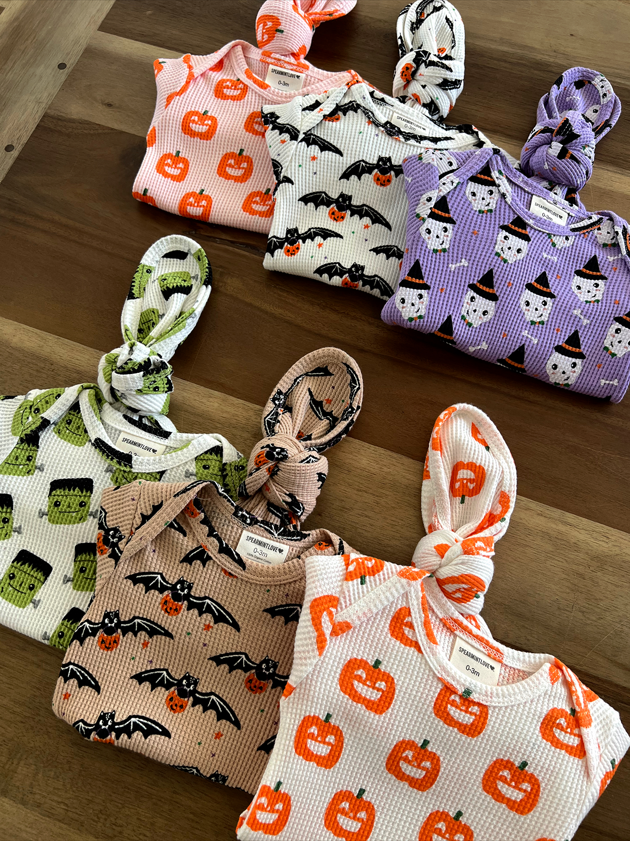Halloween-themed infant clothes with pumpkin, bat, and ghost patterns in soft fabric, displayed on a wooden surface.