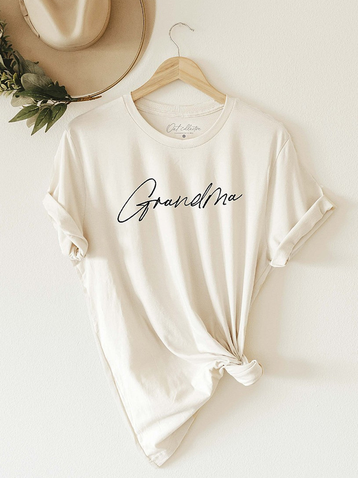 Cream t-shirt with "GrandMa" printed in cursive, knotted at the waist, displayed on a hanger against a wall.