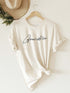 Cream t-shirt with "GrandMa" printed in cursive, knotted at the waist, displayed on a hanger against a wall.