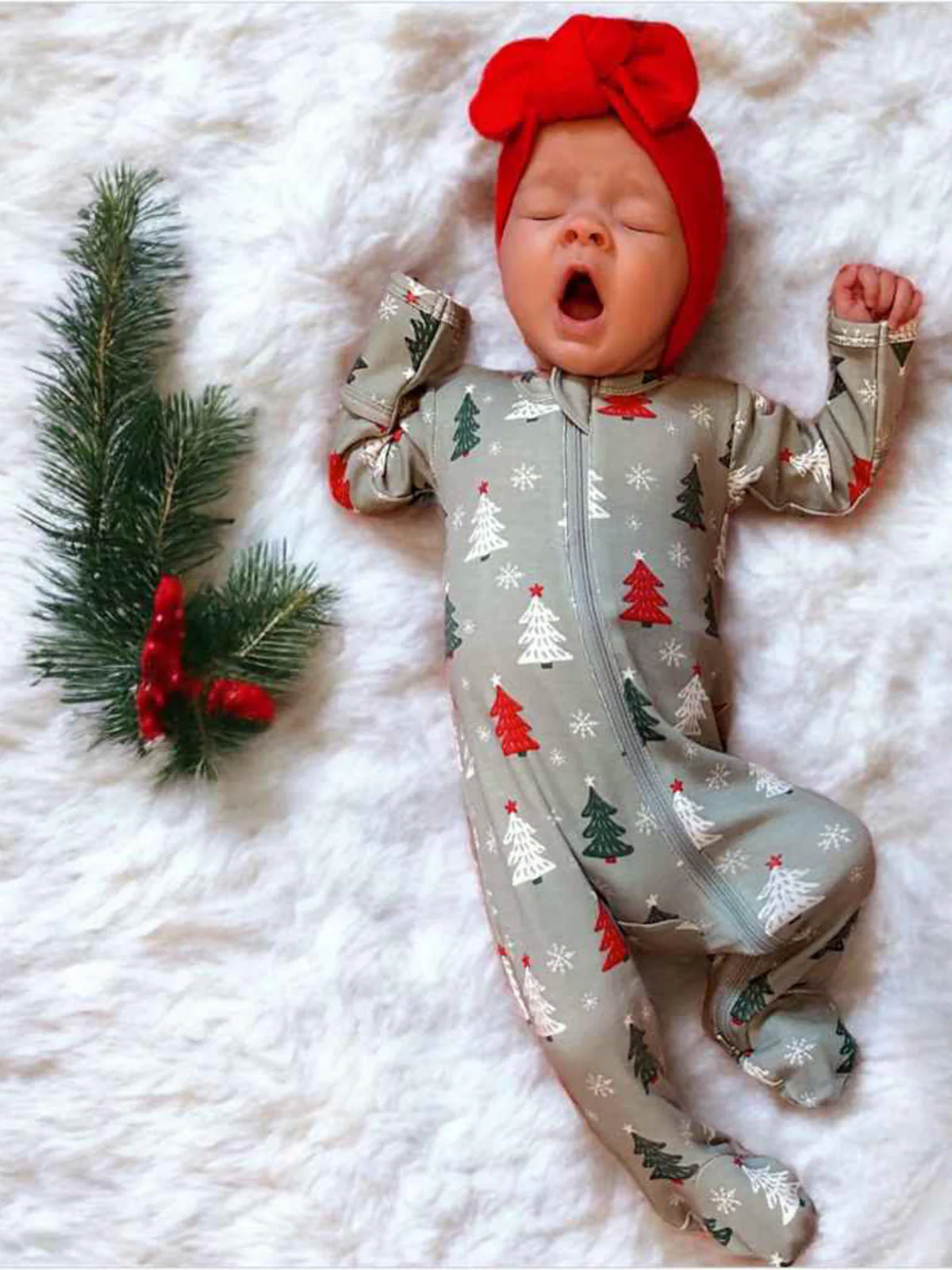 Baby in festive pajamas yawning, surrounded by Christmas decorations on a fluffy white blanket.