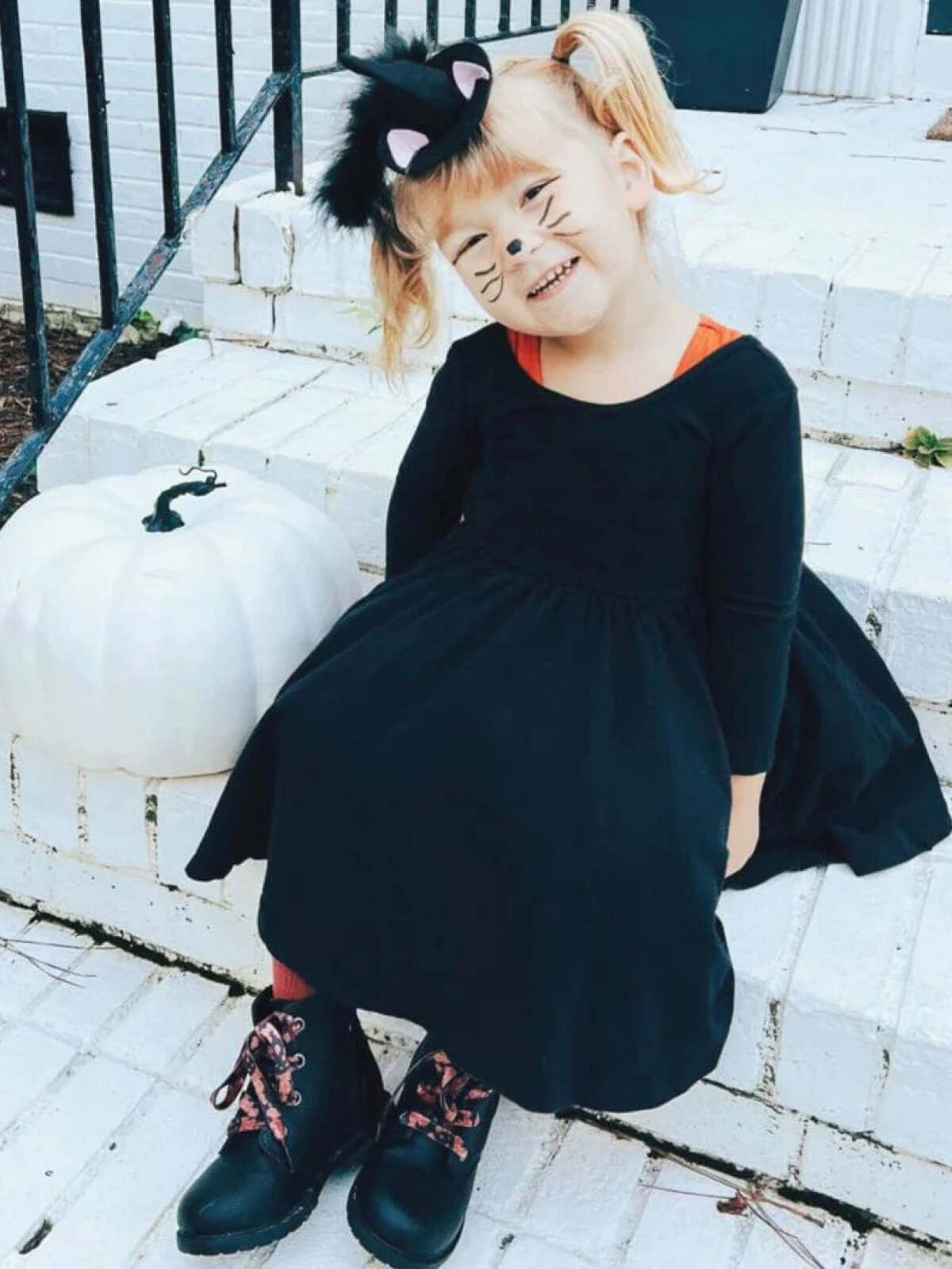 Smiling child in a black cat costume sitting by a white pumpkin on a porch.