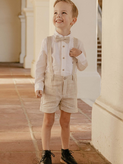 Smiling boy in a stylish outfit with bowtie and suspenders, standing on a tiled floor in a bright setting.