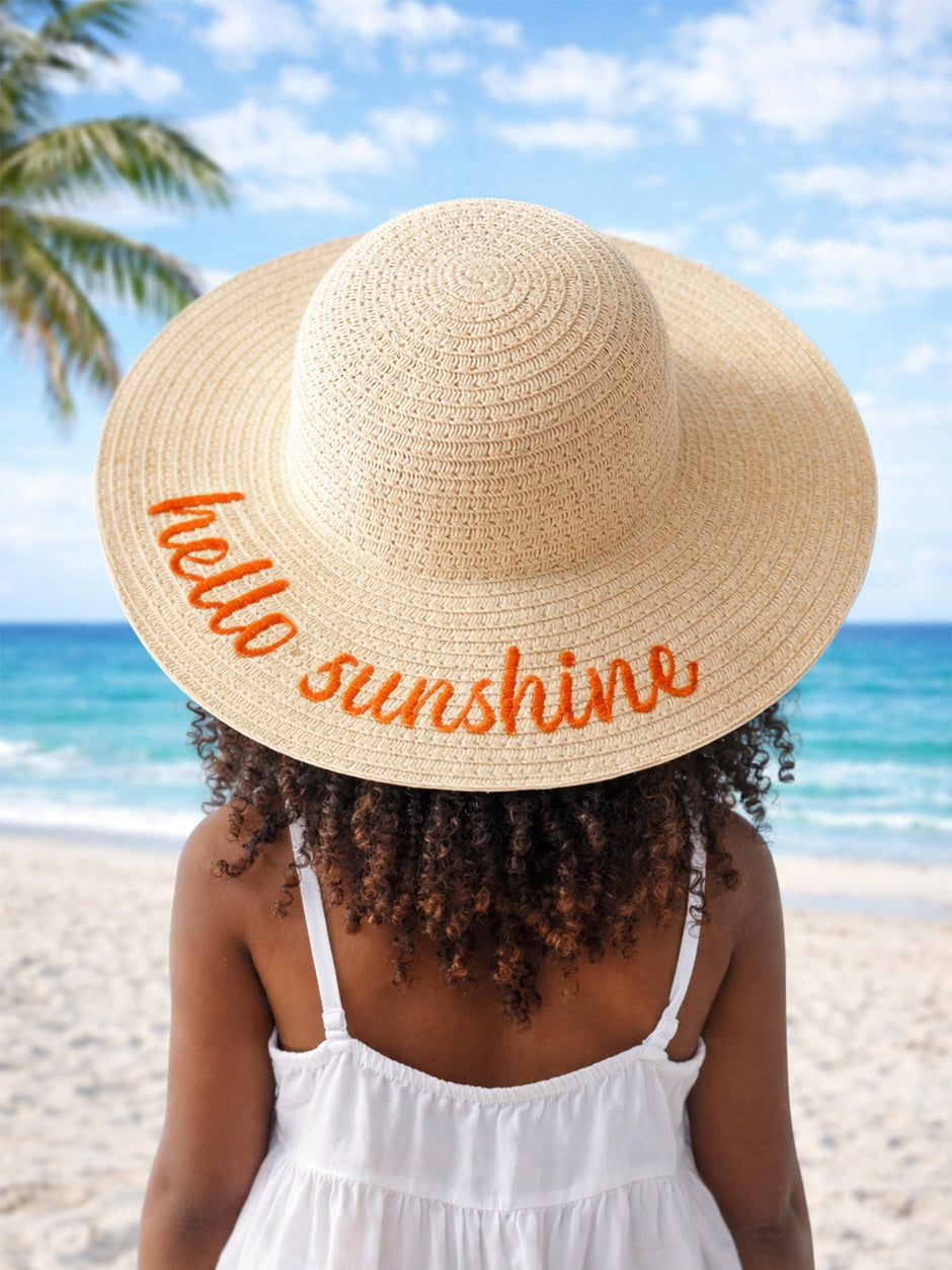 Girl wearing a large straw hat that says "hello sunshine," with a beach and palm trees in the background.