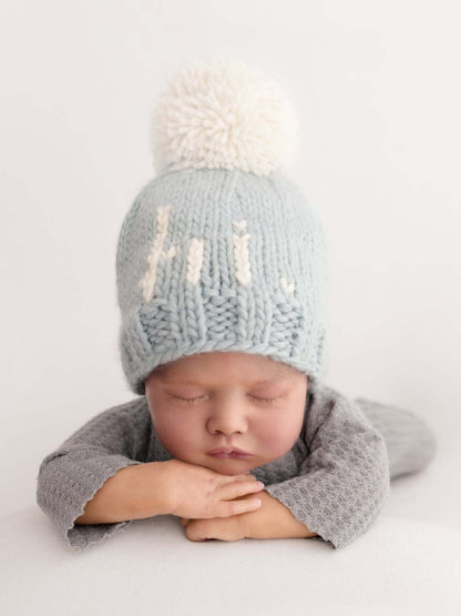 Newborn doll wearing a soft blue knit hat with a white pompom, resting peacefully with arms crossed.