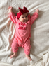 Baby lying on a white blanket, smiling, wearing a pink outfit with “HOLLY JOLLY” text and a red bow headband.
