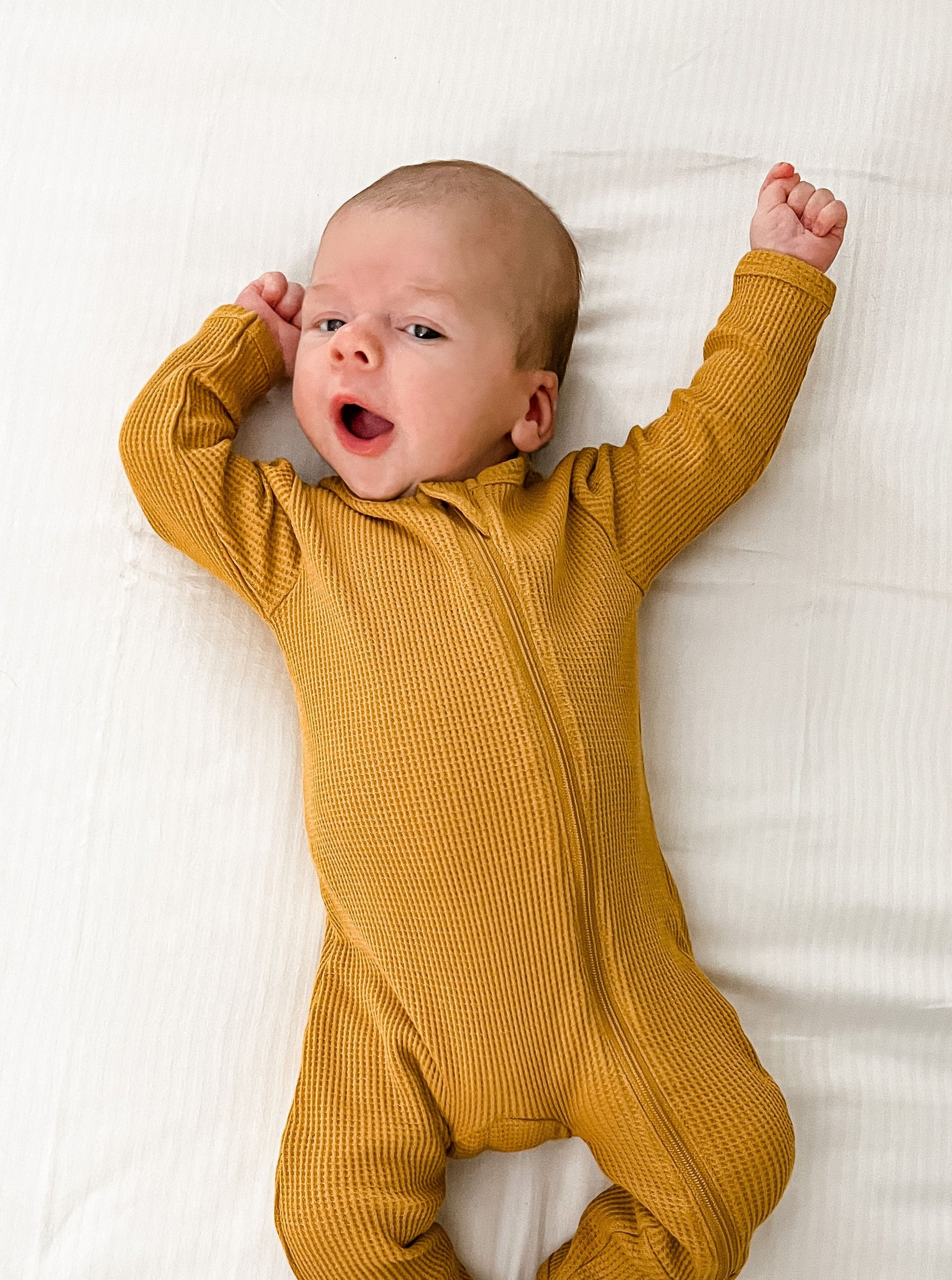 Baby in a yellow onesie yawning with arms stretched, lying on a white surface.