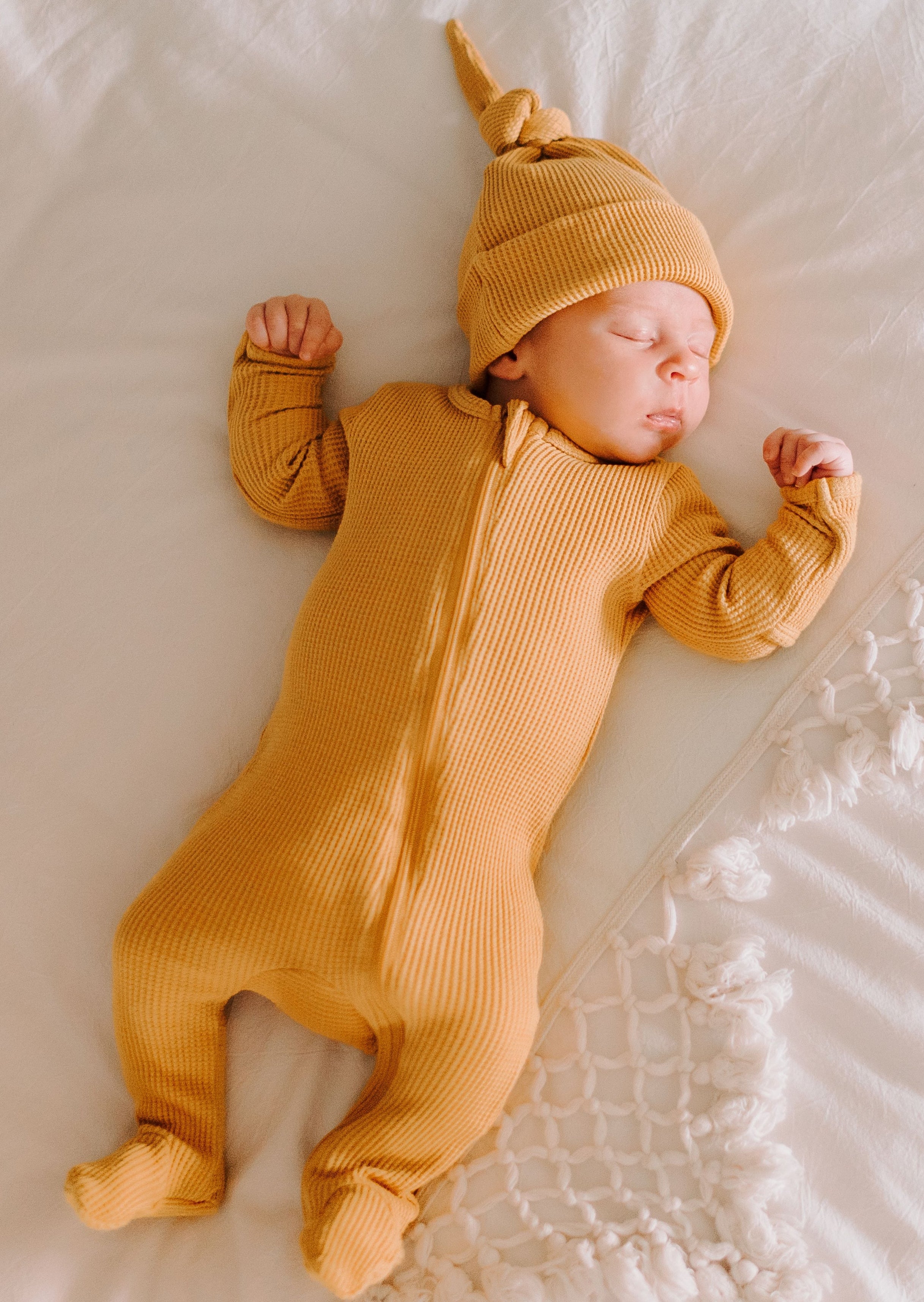 Sleeping baby in a yellow ribbed onesie and matching hat, resting on a soft white blanket.