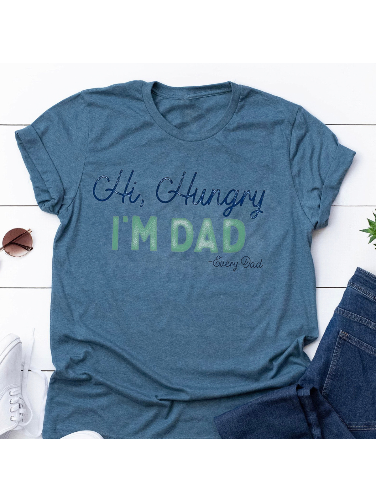 Blue t-shirt with "Hi, Hungry, I'm Dad" design, styled with jeans and sneakers on a wooden background.